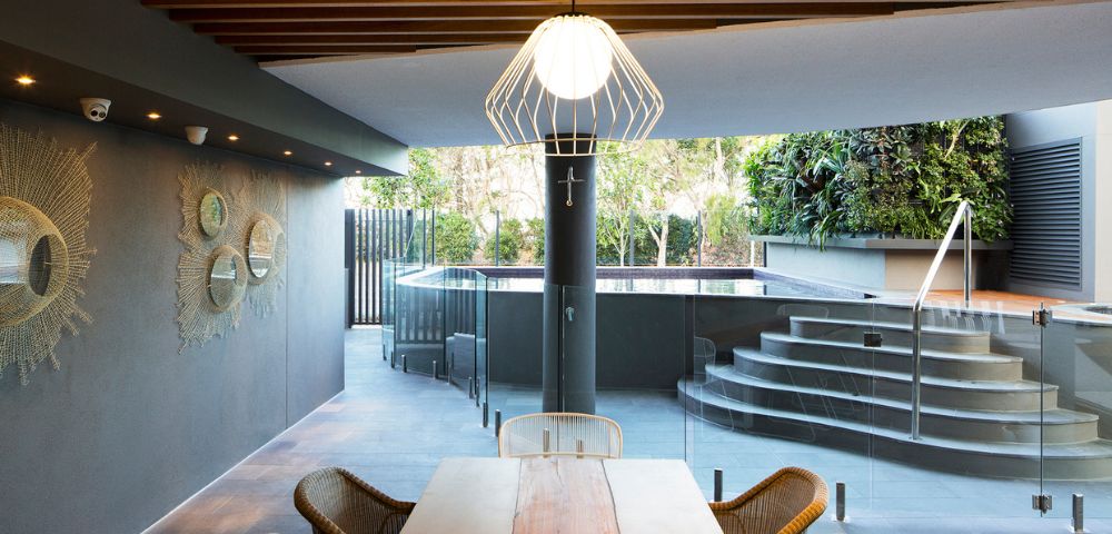 Stylish outdoor dining area with wooden table and wicker chairs. A modern pendant light hangs above, leading to a sleek pool with steps and lush greenery.