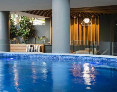 Indoor pool area with shimmering blue water in the foreground. Modern seating and lush plants by a wood-paneled wall create a serene atmosphere.