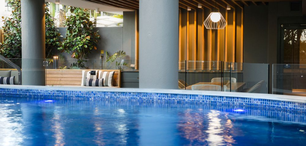 Indoor pool area with shimmering blue water in the foreground. Modern seating and lush plants by a wood-paneled wall create a serene atmosphere.