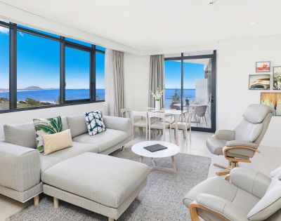 Modern, bright living room with large windows showcasing ocean views. Features a gray sectional sofa, cozy armchairs, and a dining table, creating a serene atmosphere.