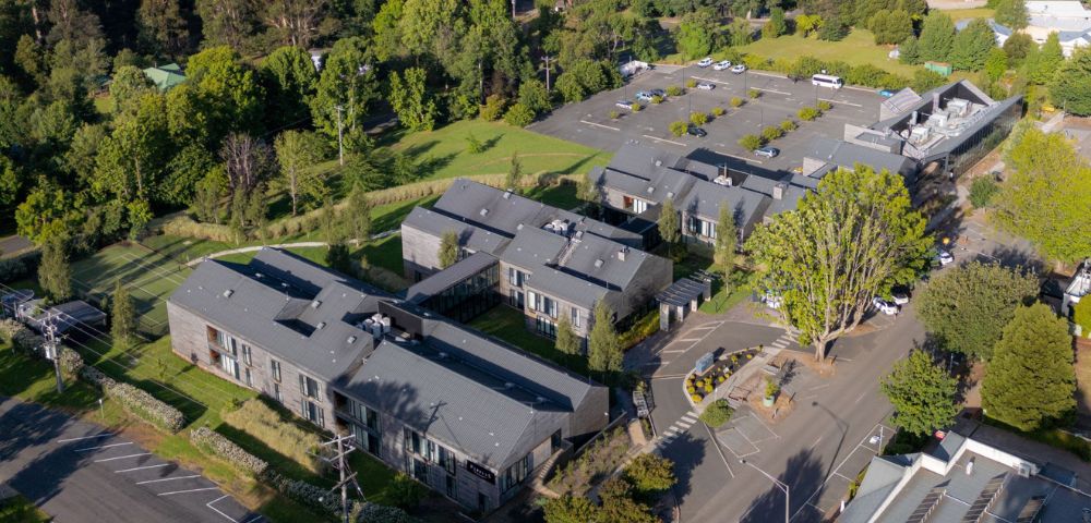 Aerial view of a modern campus with grey buildings surrounded by trees and greenery, featuring a large parking area. Tranquil and organized setting.