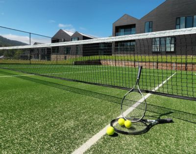 A tennis court with vibrant green grass, two racquets leaning on the net, and several tennis balls scattered. Modern buildings and a clear sky in the background.