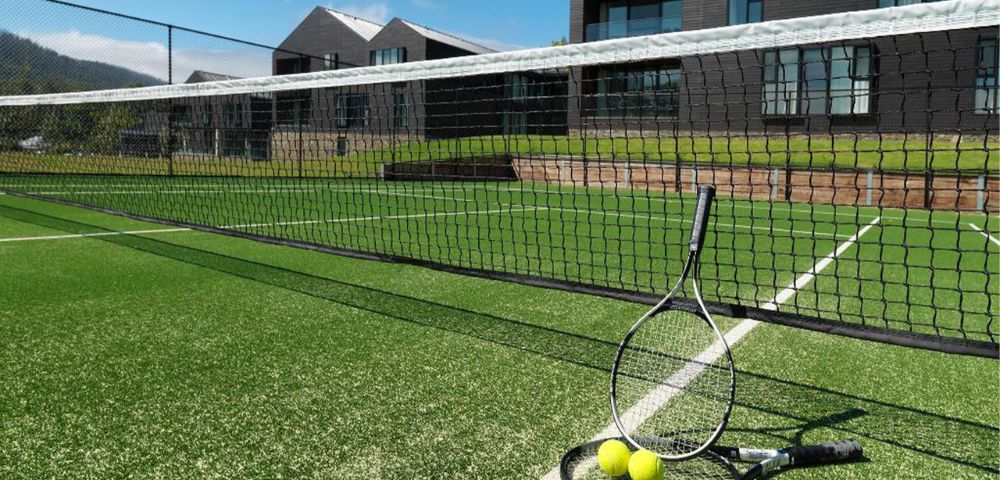 A tennis court with vibrant green grass, two racquets leaning on the net, and several tennis balls scattered. Modern buildings and a clear sky in the background.