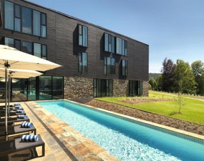 Modern building with a stone facade and large windows beside a sleek swimming pool. Lounge chairs and umbrellas line the pool, with green lawns beyond.