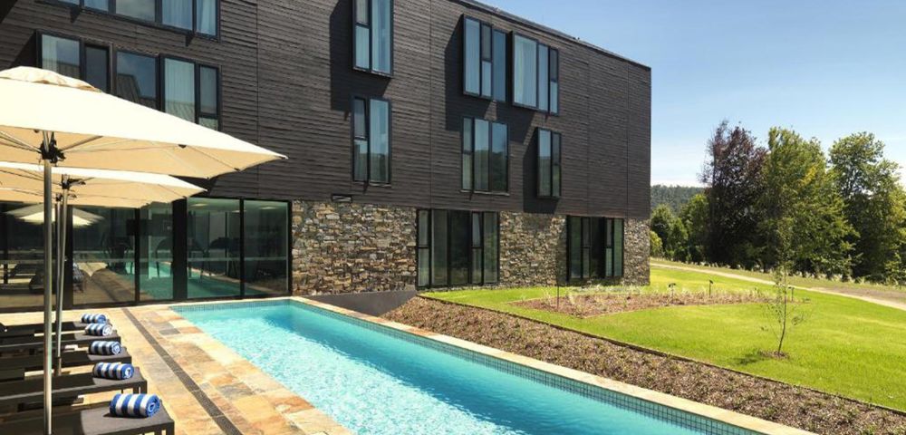 Modern building with a stone facade and large windows beside a sleek swimming pool. Lounge chairs and umbrellas line the pool, with green lawns beyond.