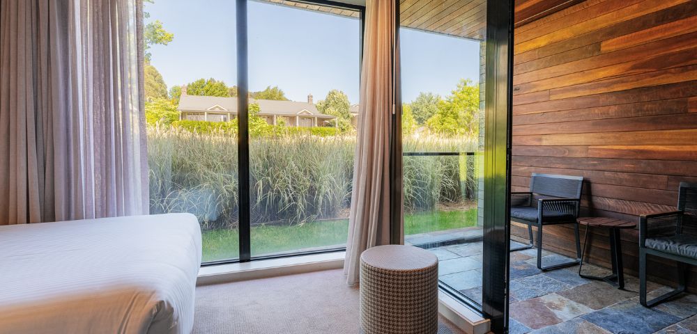 A serene bedroom with a full-length window showing lush green grass and trees. Light curtains, a white bed, and a small cylindrical ottoman add a cozy feel.