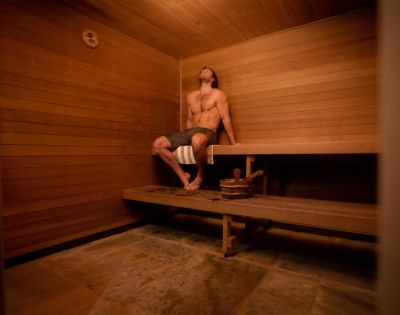 Shirtless man sitting relaxed on a wooden bench in a dimly lit sauna with wooden walls and a stone floor, creating a warm, tranquil atmosphere.