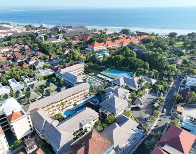 Aerial view of a coastal resort complex with red-roofed buildings, lush greenery, and swimming pools, adjacent to a sandy beach and blue ocean.