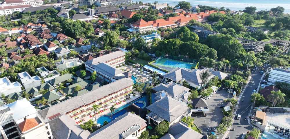 Aerial view of a coastal resort complex with red-roofed buildings, lush greenery, and swimming pools, adjacent to a sandy beach and blue ocean.