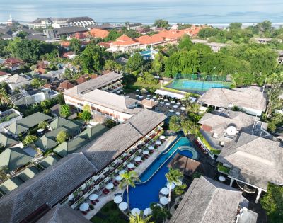 Aerial view of a tropical resort with lush greenery, a central curved pool surrounded by loungers, tennis courts, and buildings with thatched roofs. Calm ambiance.