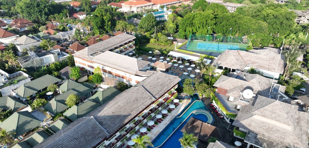 Aerial view of a tropical resort with lush greenery, a central curved pool surrounded by loungers, tennis courts, and buildings with thatched roofs. Calm ambiance.