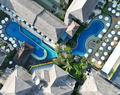 Aerial view of a tropical resort with two curvy pools surrounded by sunbeds and lush greenery. Buildings with thatched roofs frame the pools, creating a relaxing atmosphere.