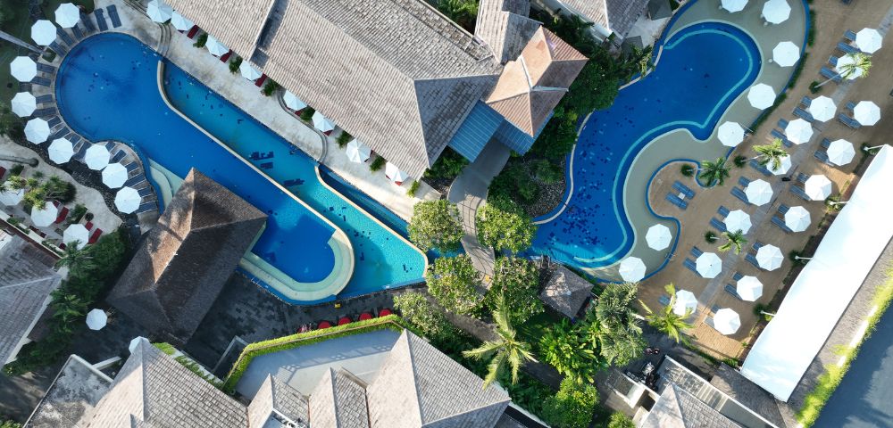 Aerial view of a tropical resort with two curvy pools surrounded by sunbeds and lush greenery. Buildings with thatched roofs frame the pools, creating a relaxing atmosphere.