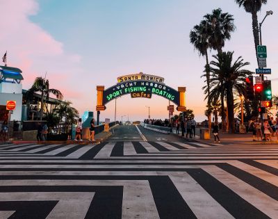 A vibrant sunset skyline frames the Santa Monica pier entrance, with colorful neon lights, palm trees, and people crossing the striped crosswalk.