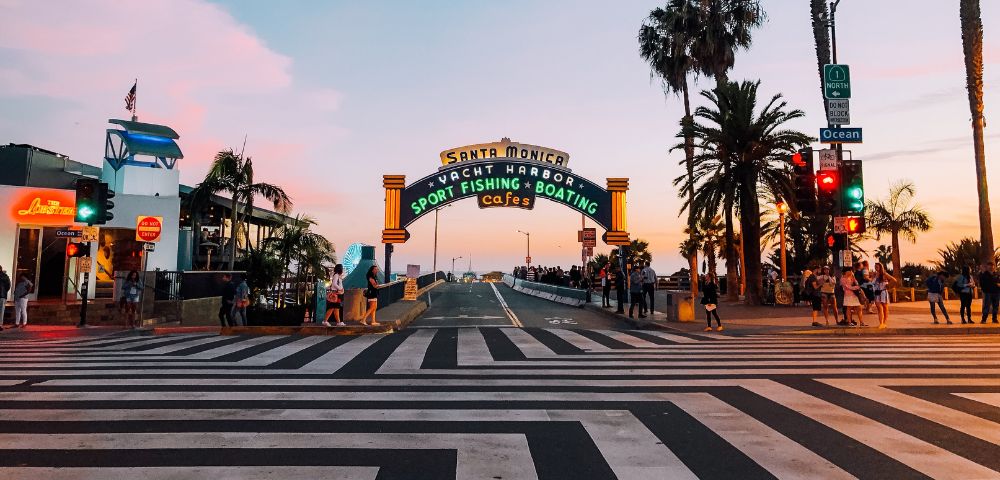 A vibrant sunset skyline frames the Santa Monica pier entrance, with colorful neon lights, palm trees, and people crossing the striped crosswalk.