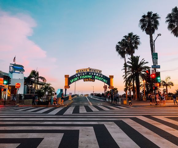 A vibrant sunset skyline frames the Santa Monica pier entrance, with colorful neon lights, palm trees, and people crossing the striped crosswalk.