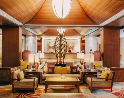 Luxurious hotel lobby with warm lighting, wooden ceiling, and elegant decor. Central seating area features plush cushions around a modern sculpture, evoking a welcoming ambiance.
