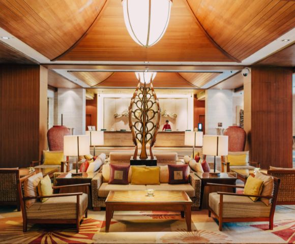 Luxurious hotel lobby with warm lighting, wooden ceiling, and elegant decor. Central seating area features plush cushions around a modern sculpture, evoking a welcoming ambiance.