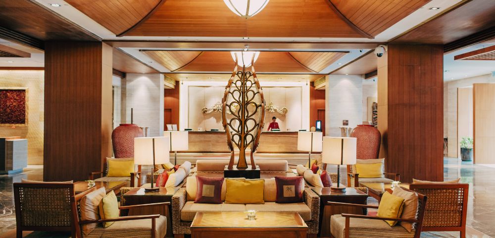 Luxurious hotel lobby with warm lighting, wooden ceiling, and elegant decor. Central seating area features plush cushions around a modern sculpture, evoking a welcoming ambiance.