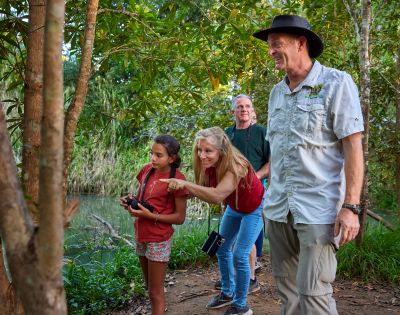 A group of four people, including a child with binoculars, explore a lush, green forest path by a river. They look curious and engaged with nature.