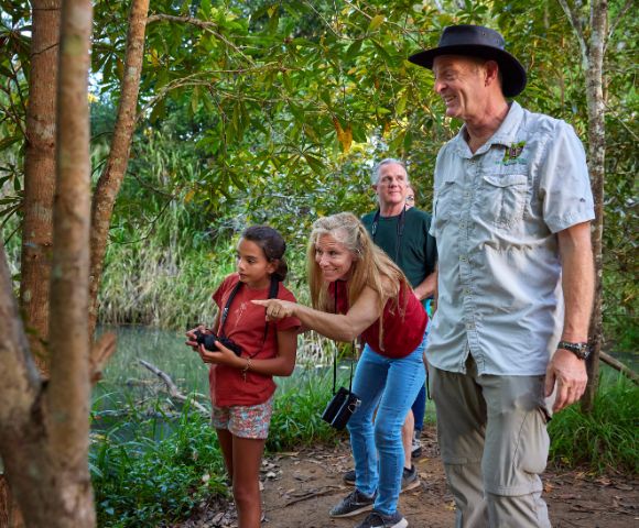 A group of four people, including a child with binoculars, explore a lush, green forest path by a river. They look curious and engaged with nature.