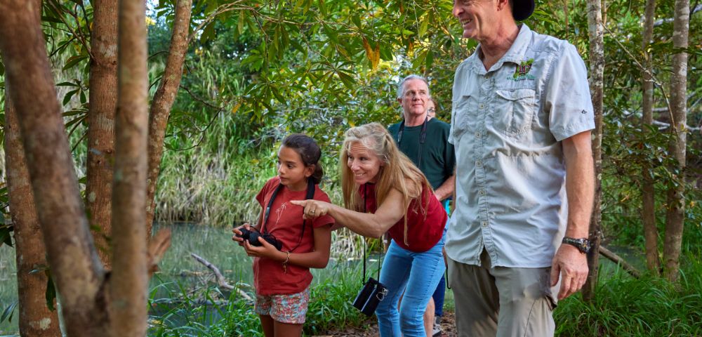 A group of four people, including a child with binoculars, explore a lush, green forest path by a river. They look curious and engaged with nature.