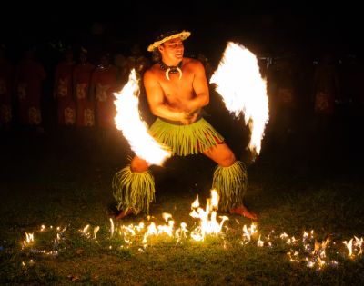 A performer in traditional attire dances with flaming torches in both hands, creating arcs of fire. The scene is vibrant and captivating under the night sky.
