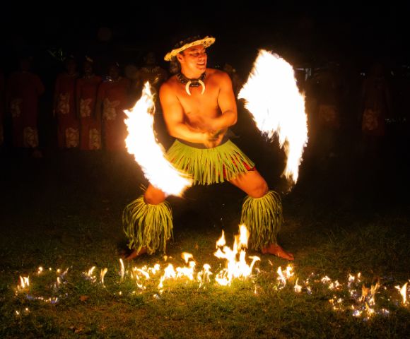 A performer in traditional attire dances with flaming torches in both hands, creating arcs of fire. The scene is vibrant and captivating under the night sky.