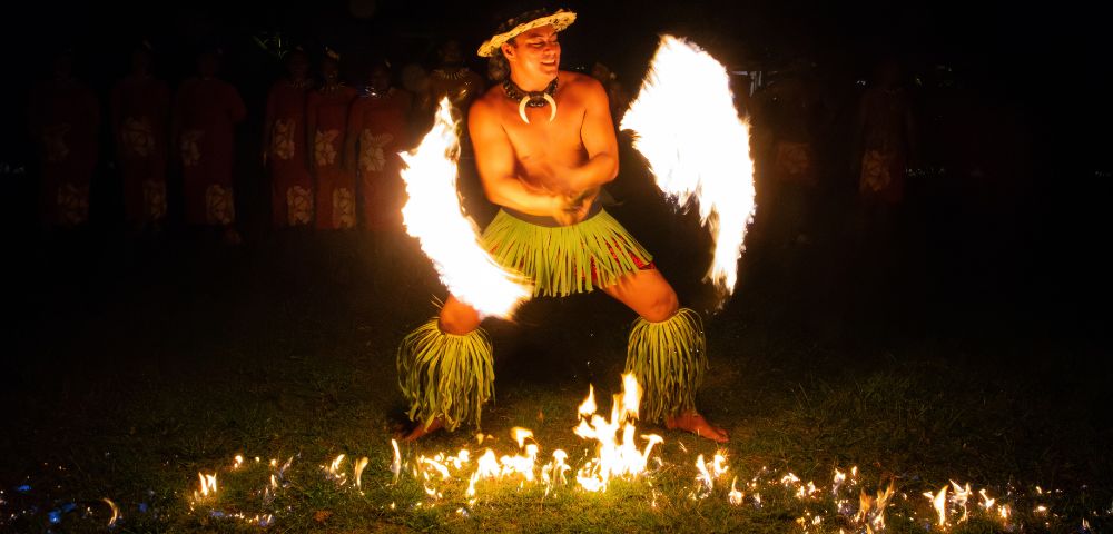 A performer in traditional attire dances with flaming torches in both hands, creating arcs of fire. The scene is vibrant and captivating under the night sky.