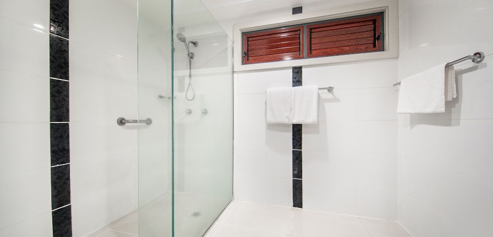Minimalist bathroom with a glass shower partition, black tile accents, white walls and floor, wooden window shutters, and neatly hung towels.