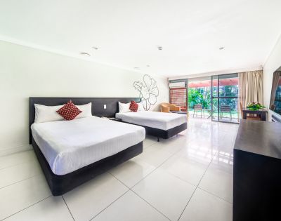 A spacious hotel room features two double beds with white linens and red accent pillows, a wall mural, and a view of a lush garden through glass doors.