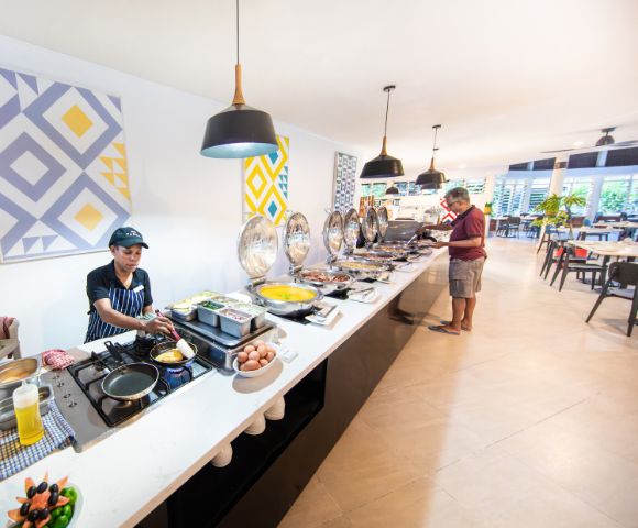 A buffet service area with a chef preparing food on a counter filled with dishes. A man is serving himself, in a bright, modern dining room.