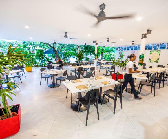 Bright, airy restaurant with white tables, black chairs, and ceiling fans. A server walks through. Green plants line the windows, creating a fresh ambiance.