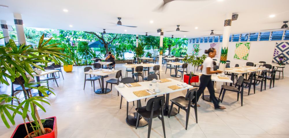 Bright, airy restaurant with white tables, black chairs, and ceiling fans. A server walks through. Green plants line the windows, creating a fresh ambiance.
