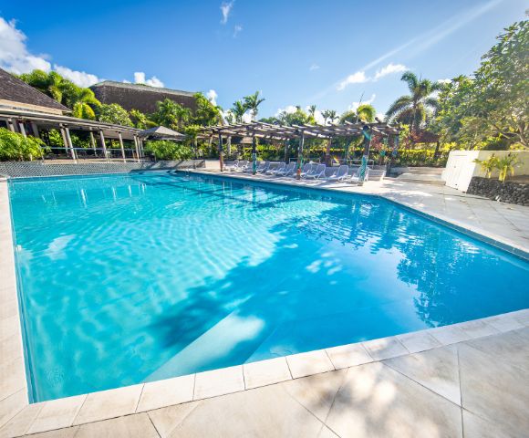 A tranquil swimming pool with clear blue water, surrounded by sun loungers and tropical palm trees, under a bright blue sky with a few clouds.