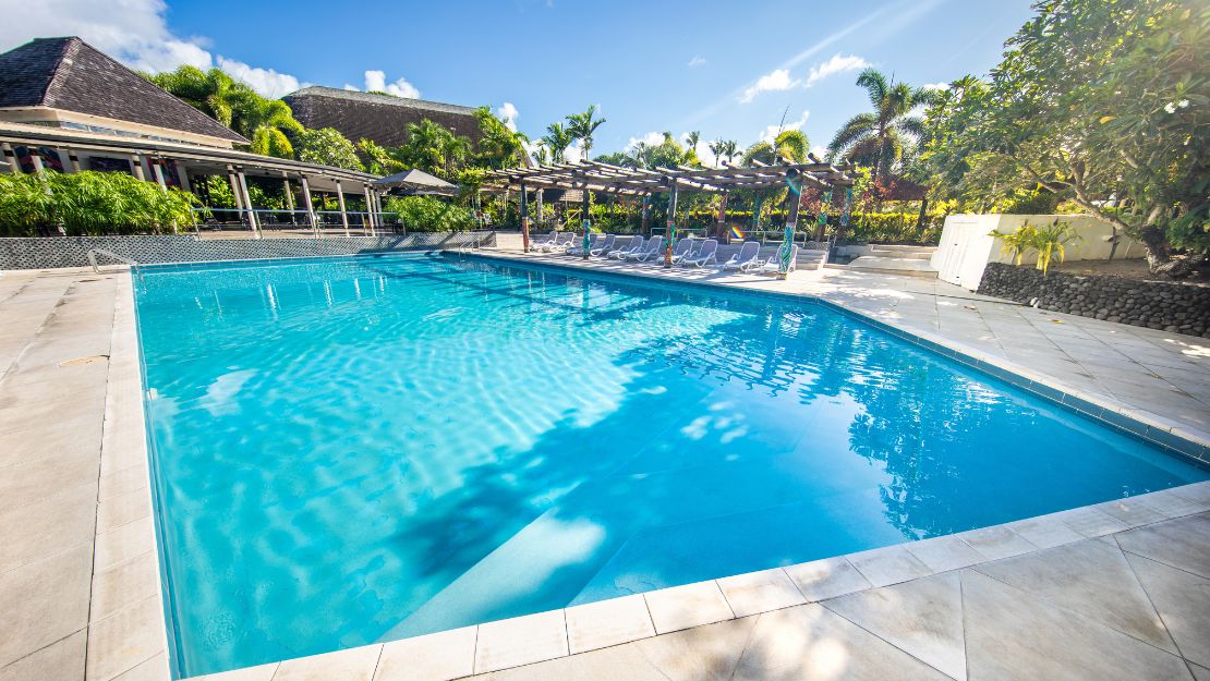 A tranquil swimming pool with clear blue water, surrounded by sun loungers and tropical palm trees, under a bright blue sky with a few clouds.