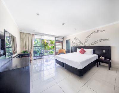 Bright, modern hotel room with a large bed, red accent pillow, and sleek furniture. Glass doors lead to a balcony with lush greenery outside. Calm ambiance.
