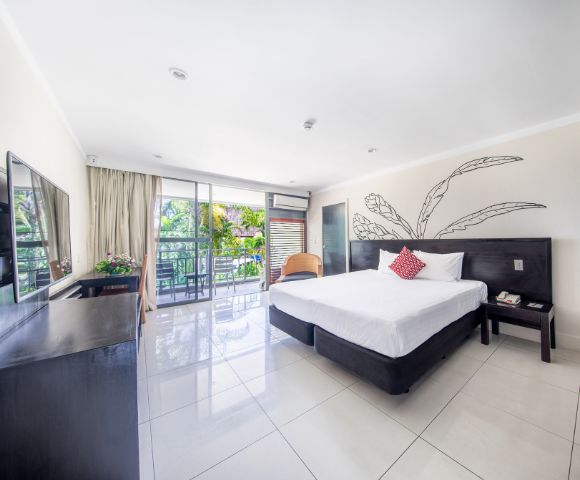 Bright, modern hotel room with a large bed, red accent pillow, and sleek furniture. Glass doors lead to a balcony with lush greenery outside. Calm ambiance.