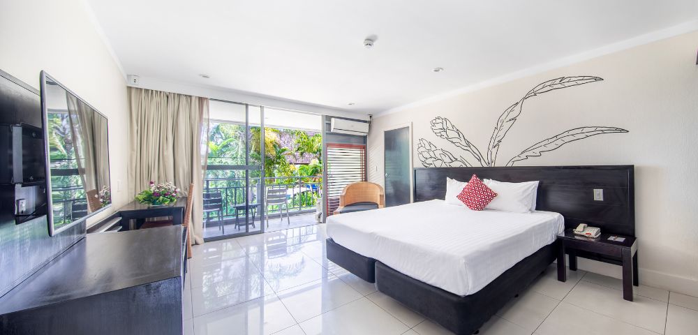 Bright, modern hotel room with a large bed, red accent pillow, and sleek furniture. Glass doors lead to a balcony with lush greenery outside. Calm ambiance.