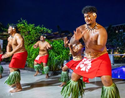 Four men in traditional Polynesian attire dance joyfully, wearing red skirts and grass anklets. The scene is vibrant, set against a lush, tropical backdrop at night.