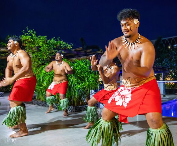 Four men in traditional Polynesian attire dance joyfully, wearing red skirts and grass anklets. The scene is vibrant, set against a lush, tropical backdrop at night.