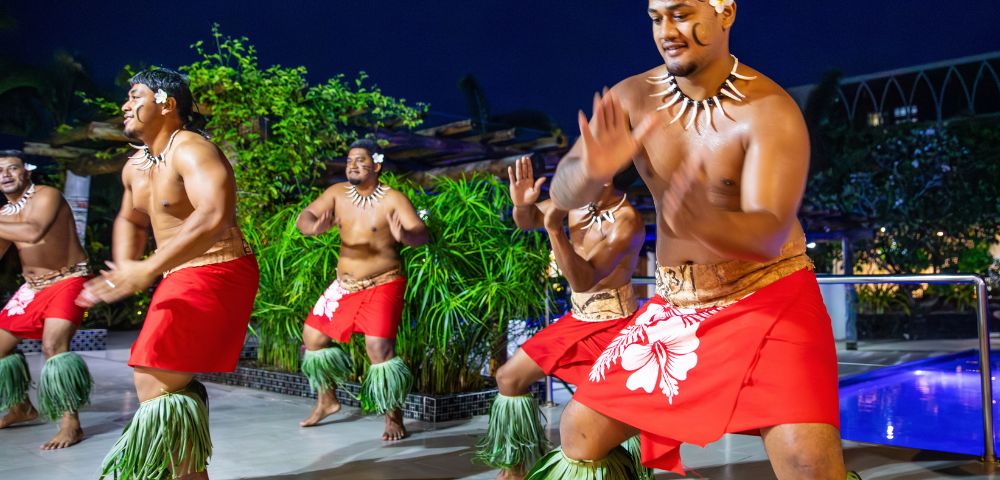 Four men in traditional Polynesian attire dance joyfully, wearing red skirts and grass anklets. The scene is vibrant, set against a lush, tropical backdrop at night.