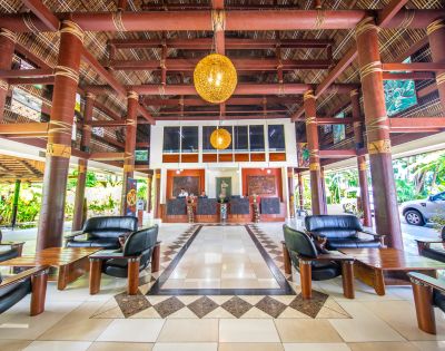 Spacious tropical hotel lobby with bamboo columns, geometric tile design, black leather seating, and warm wooden accents. Casual, inviting ambiance.