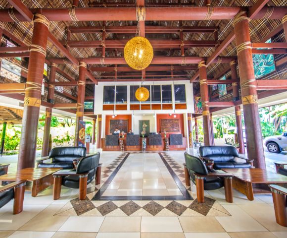 Spacious tropical hotel lobby with bamboo columns, geometric tile design, black leather seating, and warm wooden accents. Casual, inviting ambiance.