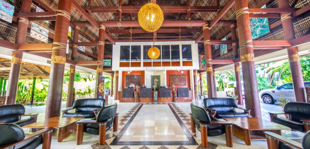 Spacious tropical hotel lobby with bamboo columns, geometric tile design, black leather seating, and warm wooden accents. Casual, inviting ambiance.