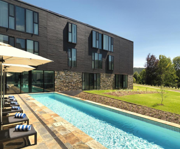 A modern hotel with a sleek, dark facade stands beside a narrow turquoise swimming pool. Sun loungers with striped towels line the poolside, under large umbrellas.