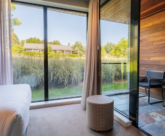 Modern bedroom with large glass doors opening to a patio. Bright natural light, lush greenery outside, wooden accents, serene and cozy atmosphere.