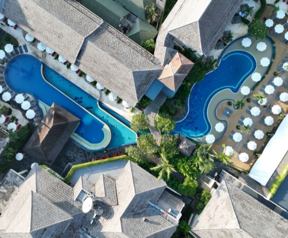 Aerial view of a resort with two curvy blue pools surrounded by sun umbrellas and loungers. Gray-roofed buildings enclose the pools amidst lush greenery.