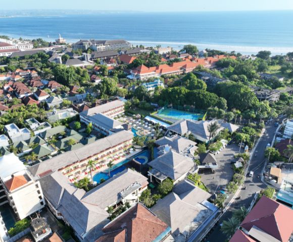 Aerial view of a coastal hotel resort surrounded by lush greenery and red-roofed buildings near a beach. The ocean is visible in the background.