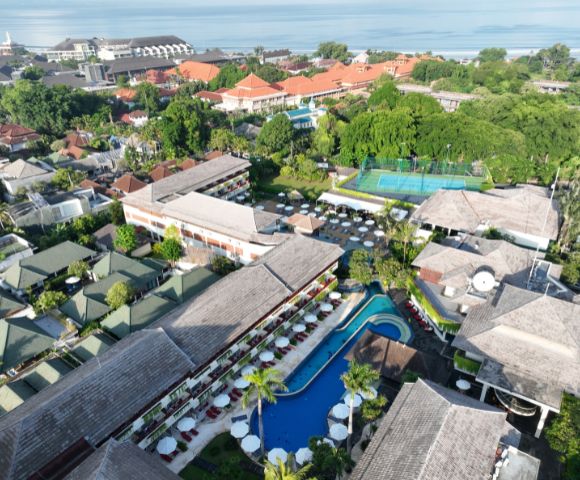 Aerial view of a tropical resort with blue lagoon-style pool, surrounded by lush greenery and traditional red-roofed buildings, creating a serene ambiance.